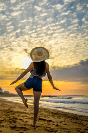 Joyful woman dances barefoot on sand beach on beautiful dawn sky background. Back viewの写真素材
