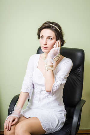 Beautiful business woman in white dress sitting on the chair and holding mobile phone over light...