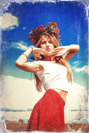Portrait of amazing girl in wildflower wreath and red skirt in countryside. Young woman with raised hands looking at camera on blurred overexposed outdoor background.の写真素材