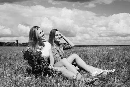 Black and white portrait of attractive two young ladies sitting on summer meadow. Pretty girls with flower bouquet on retro filtered summer countryside background.の写真素材
