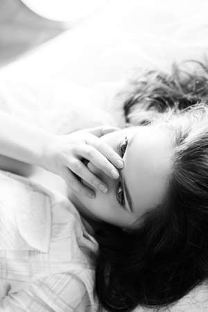 Black and white photography of young woman lying in bed. Charming girl gently looking up on blurred indoor background.の写真素材
