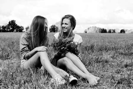Black and white photography of two charming girls sitting on grass with wildflowers. Young woman talking and happy smiling on summer countryside background.の写真素材