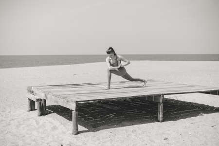 Black and white photography of morning training on the beach. Beautiful female doing workout on sunny outdoors background. Healthy lifestyle exercises. Back side view of perfect fit shape body.の写真素材
