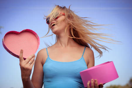 Joyful excited lovely woman holding heart shaped gift box in hands. Happy smiling surprised young lady over light blue sky background. Excitement beauty celebrating momentの写真素材