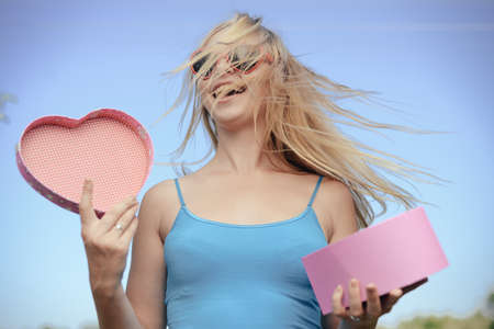 Joyful excited lovely woman holding heart shaped gift box in hands. Happy smiling surprised young lady over light blue sky background. Excitement beauty celebrating momentの写真素材