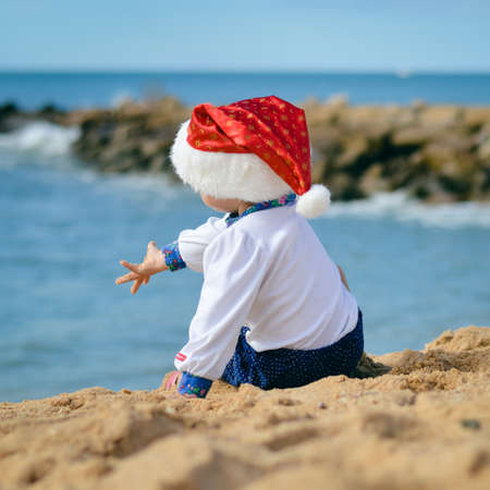 Child in Christmas Santa Claus hat on ocean tropical sandy beach seasonal travel vacation, blue sky background outdoors. Back side viewの写真素材