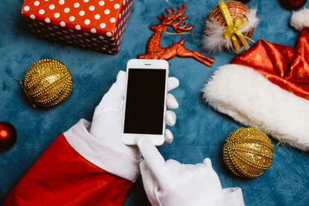 Close up on Santa holding use smart phone, mockup copyspace Christmas eve. Business wireless application for shopping delivery. Portable camera online internet technology. Hat, toy, decor happy eventの写真素材