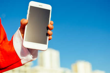Happy cute Santa child holding mobile smartphone showing blank display for your text message on light blue sky background. Happiness shopping time and electronic education lifestyle. Close up photoの写真素材
