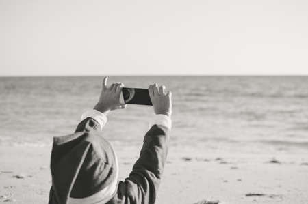 Child boy Santa Claus using mobile phone on ocean beach outdoors background, back view photo. Presents and gifts festive time new year holiday seasonの写真素材