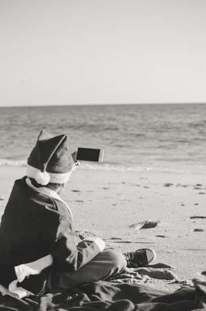 Child boy Santa Claus using mobile phone on ocean beach outdoors background, back view photo. Presents and gifts festive time new year holiday seasonの写真素材