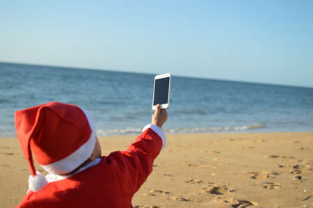 Happy cute Santa child holding mobile smartphone showing blank display for your text message on light blue sky background. Happiness shopping time and electronic education lifestyle. Close up photoの写真素材