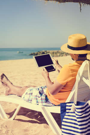 Businessman holding in hands using mobile tablet pc computer on sunny beach outdoors holiday. Back view mock up travel vacation backgroundの写真素材