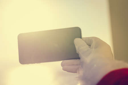 Busy Santa Claus holding mobile smartphone ready for Christmas over sun light blue sky. Close up view on person showing mockup display app. Happy sunny seasonal festive time photographyの写真素材