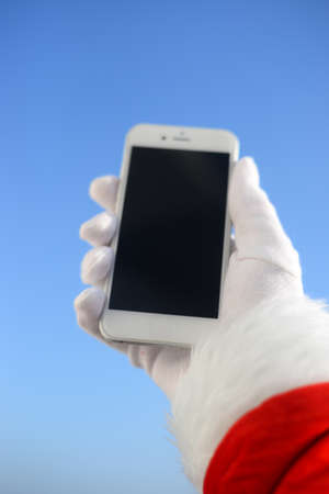 Busy Santa Claus holding mobile smartphone ready for Christmas over sun light blue sky. Close up view on person showing mockup display app. Happy sunny seasonal festive time photographyの写真素材
