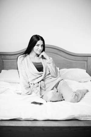 Black and white photography of beautiful young lady having fun relaxing with mobile phone in bed over light backgroundの写真素材