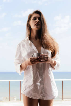 Cheerful beautiful woman enjoying healthy relax lifestyle drinking hot beverage looking out window having positive day and bright sunny oceanfront outdoors background. Travel relaxation destinationの写真素材