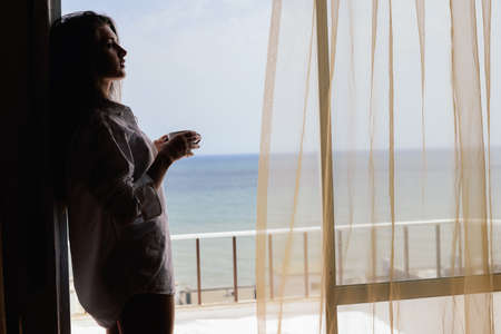 Cheerful beautiful woman enjoying healthy relax lifestyle drinking hot beverage looking out window having positive day and bright sunny oceanfront outdoors background. Travel relaxation destinationの写真素材