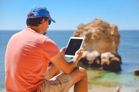 Back side view of person sitting holding tablet computer on ocean sunny natural background. Traveler having fun on international travelling lifestyle, technology social communication, amazing natureの写真素材