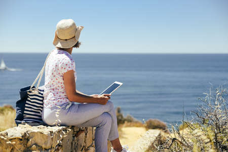 Back side view of person sitting holding tablet computer on ocean sunny natural background. Traveler...