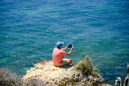 Back side view of person sitting holding tablet computer on ocean sunny natural background. Traveler having fun on international travelling lifestyle, technology social communication, amazing natureの写真素材