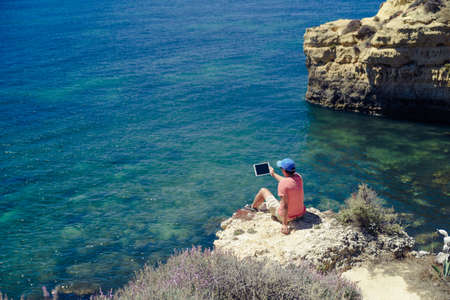 Back side view of person sitting holding tablet computer on ocean sunny natural background. Traveler having fun on international travelling lifestyle, technology social communication, amazing natureの写真素材