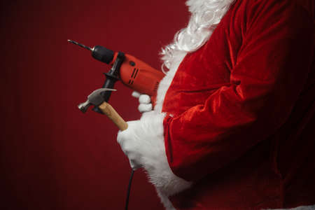 Santa Claus holding hammer and drill in hands busy preparing decoration. Closeup view of building creative ideas, taking job. Happy Christmas and New Year time renovation design backgroundの写真素材
