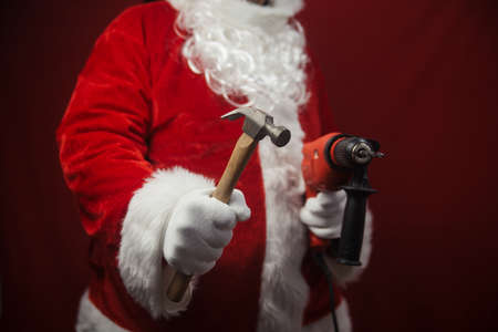 Santa Claus holding hammer and drill in hands busy preparing decoration. Closeup view of building creative ideas, taking job. Happy Christmas and New Year time renovation design backgroundの写真素材