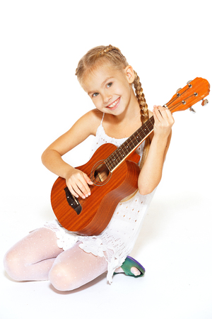 A beautiful little girl with a charming smile in a white dress with a guitarの写真素材