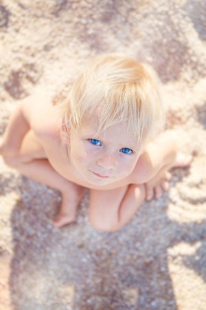 Little boy with blue eyes and white hair sitting on white sand and look up at the cameraの写真素材