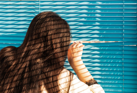 Rear view - a woman with long hair peeks out the window through the curtains of blue blindsの写真素材