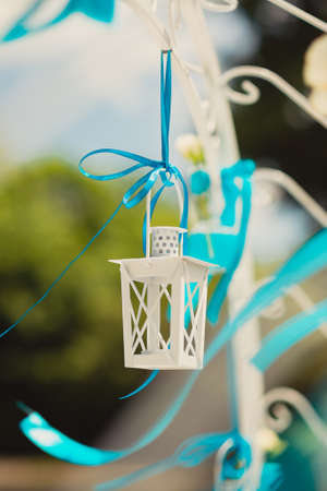 Decorative white lantern for candles on decorative white arch on a blue ribbon tied on a background of green leaves and blue skyの写真素材