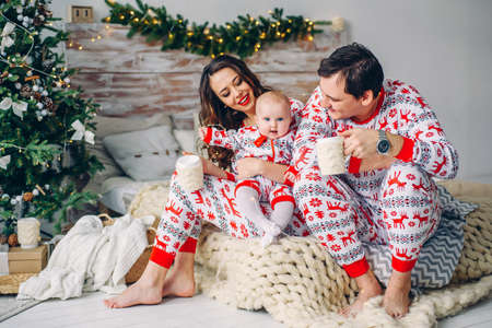 Happy parents with a baby, sitting on a bed with cups of delicious coffee in a decorated Christmas room. Parents with their child together near Christmas tree indoors.の写真素材
