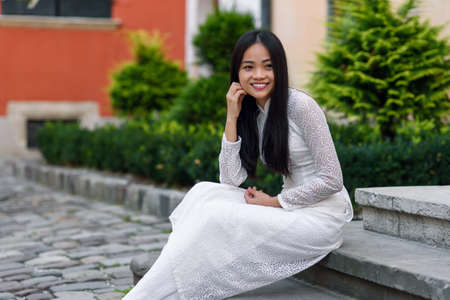 Close up portrait of beautiful asian girl dressed in Ao Dai white dress sitting on the stairs in courtyard.の写真素材