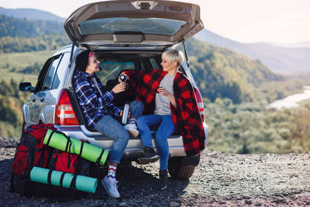 Traveling, tourism and friendship concept. Young friends travelers sitting on trunk of car and drinks hot tea. Two happy hipster girls enjoying on vacation in mountains.の写真素材
