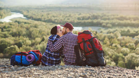 Young couple with backpacks traveling together in mountains. Happy hipster man kissing his girlfriend on the top of mountain at sunset backdrop. Traveling, tourism and relationships concept.の写真素材