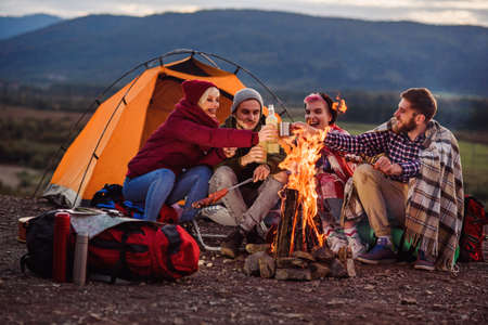 Company of young friends having a picnic by the mountains, they are chatting, laughing, drinking energy drink and bake sausages on bonfire. Concept of happiness, youth and enjoyment of life.の写真素材