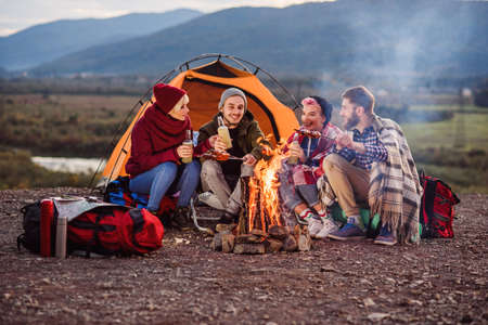 Company of young friends having a picnic by the mountains, they are chatting, laughing, drinking energy drink and bake sausages on bonfire. Concept of happiness, youth and enjoyment of life.の写真素材