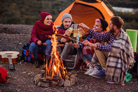 Company of young friends having a picnic by the mountains, they are chatting, laughing, drinking energy drink and bake sausages on bonfire. Concept of happiness, youth and enjoyment of life.の写真素材