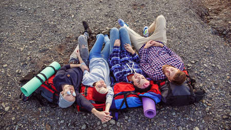 Happy friends traveling together in mountains. Happy hipster travelers with backpacks lying on the top of mountain at sunset backdrop and looking to the camera. Traveling friendship concept. Top view.の写真素材