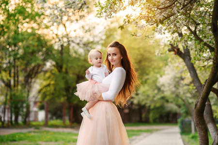 Mother holds her little daughter in her arms among blooming trees. Mom and her little baby weared pink family look dress.の写真素材