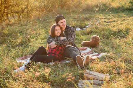 Young man hugs tenderly his pretty girlfriend. Young couple sits on the plaid at green lawn at sunset.の写真素材