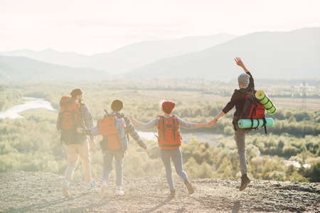 View from back of four hipster friends with travel backpack holding hands and running forward to the mountain on sunset background.の写真素材