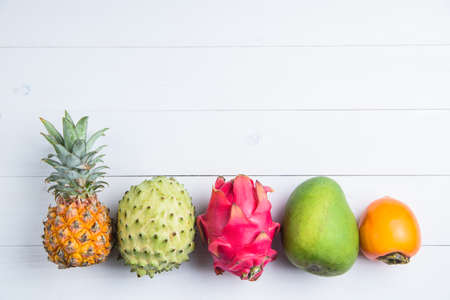Exotic fresh summer fruits on white background. Dragon fruit, pineapple, persimmon, mango, annona cherimola flat lay with free copy space.の写真素材