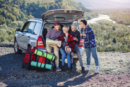 Four caucasian friends having rest after long trip while sitting in the trunk of the car, young woman pouring tea from the thermos, cute man using map.の写真素材