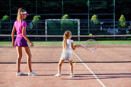 Little girl in white uniform hitting ball under net during tennis training with coach. Attractive female coach making exercises with little tennis player on the outdoor court at sunset.の写真素材