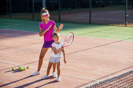 Little girl in white uniform hitting ball under net during tennis training with coach. Attractive female coach making exercises with little tennis player on the outdoor court at sunset.の写真素材