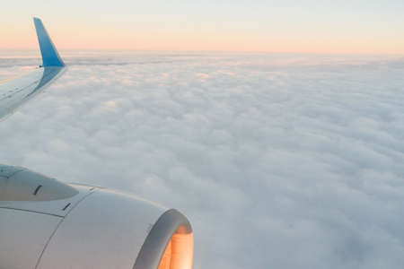 The view from the airplanes illuminator to the wing, airplane turbine and fluffy clouds at sunrise. Flying over the cloudsの写真素材