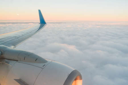 The view from the airplanes illuminator to the wing, airplane turbine and fluffy clouds at sunrise. Flying over the cloudsの写真素材