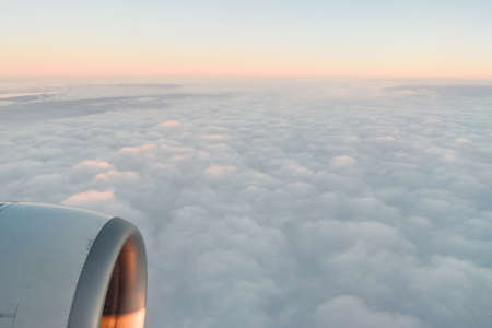 The view from the airplanes illuminator to the wing, airplane turbine and fluffy clouds at sunrise. Flying over the cloudsの写真素材