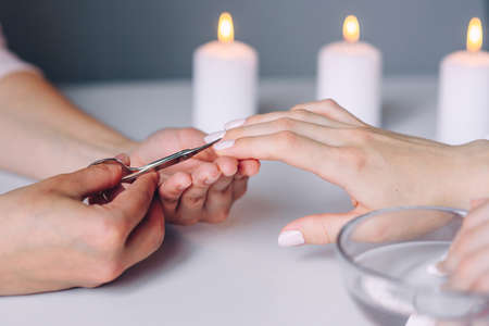 Manicurist hands uses metal cosmetic nail scissors for cutting cuticle from female clients nails. Woman hand receiving manicure procedure with nail care tool.の写真素材
